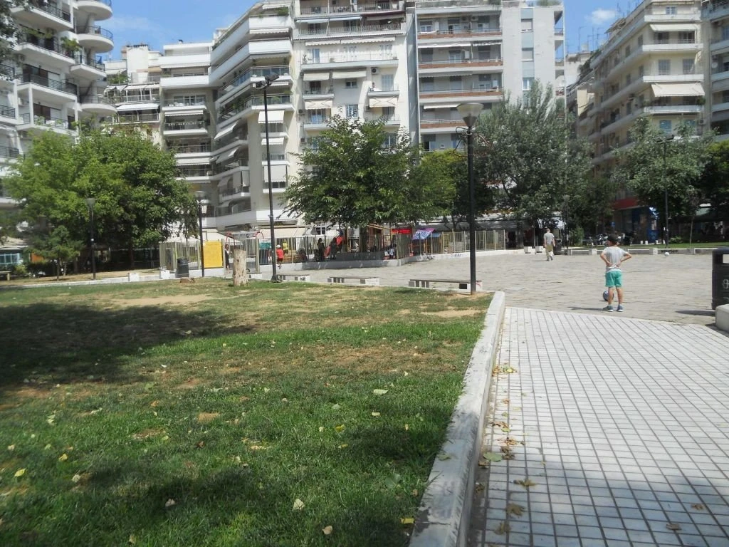 Ανατολική πλευρά του παρτεριού που θα εγκατασταθεί η νέα παιδική χαρά. Φαίνονται οι κομμένοι κορμοί, τα παλιά φωτιστικά και παγκάκια και η υφιστάμενη παιδική χαρά.